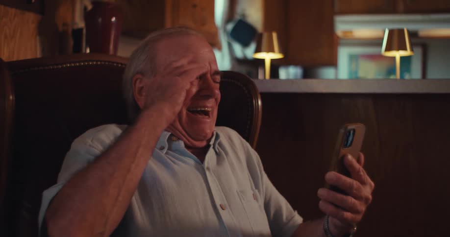The video shows a man with a blue veteran cap and a white polo shirt laughing heartily while holding a smartphone he is seated in a cozy dimly lit room with warm lighting from lamps in the background