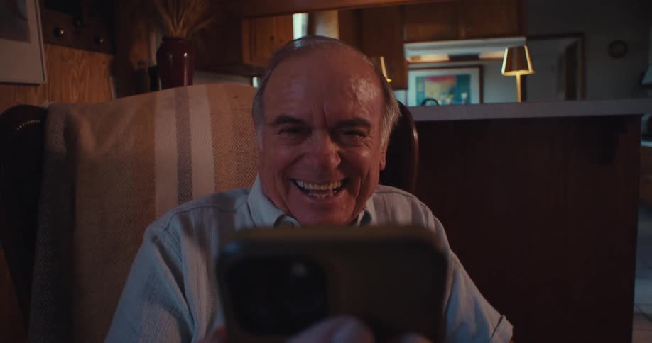An elderly man is sitting on a chair in his living room holding a phone in his hand and laughing as he looks at the screen