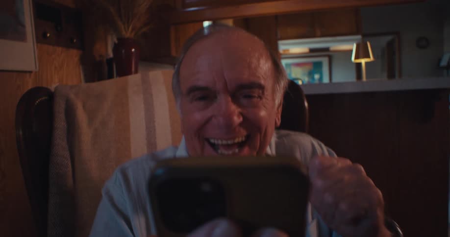 The video shows an elderly man sitting in a chair holding a smartphone and looking directly at the camera he appears to be reacting with surprise and then bursts into laughter possibly during a video call or while viewing content on his phone