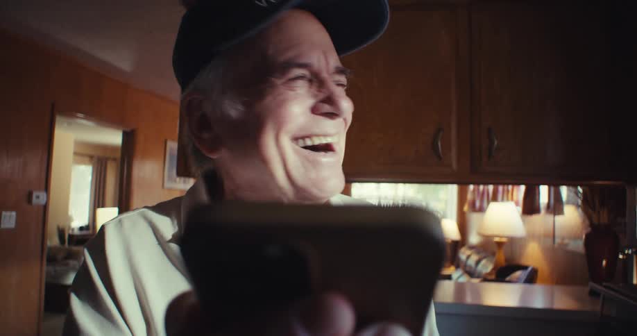 Elderly man wearing a cap laughing while holding a smartphone in front of him