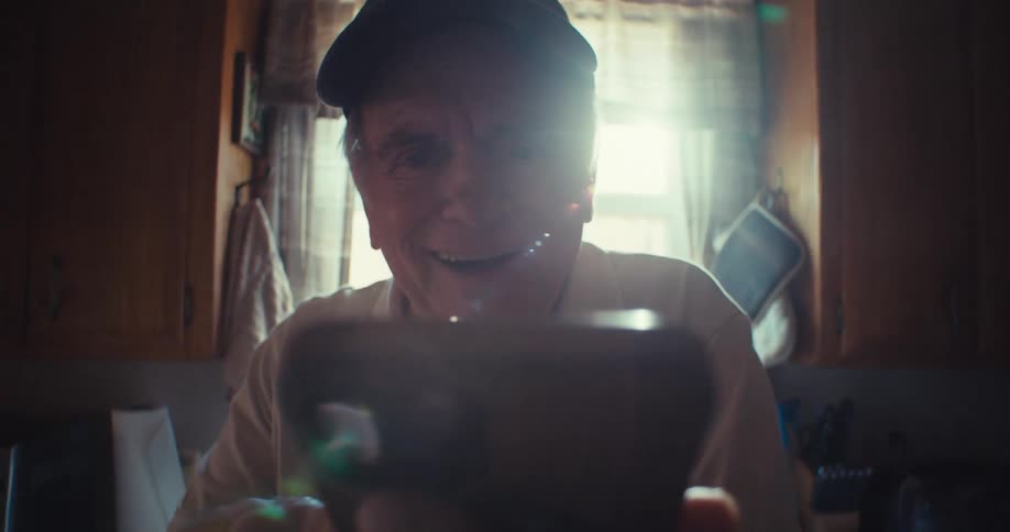 The video features a blurred closeup of an elderly man holding a smartphone with a veteran baseball cap visible he appears to be talking and laughing while looking at the screen with sunlight creating a glare in the background