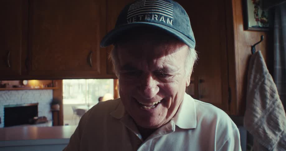 The video features an elderly man wearing a veteran cap and a white polo shirt standing in a kitchen with wooden cabinets he is looking at his smartphone and smiling suggesting he is engaged in a pleasant interaction or viewing something amusing