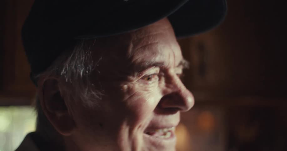 An elderly man indoors looking excited holding his phone and talking while wearing a baseball hat