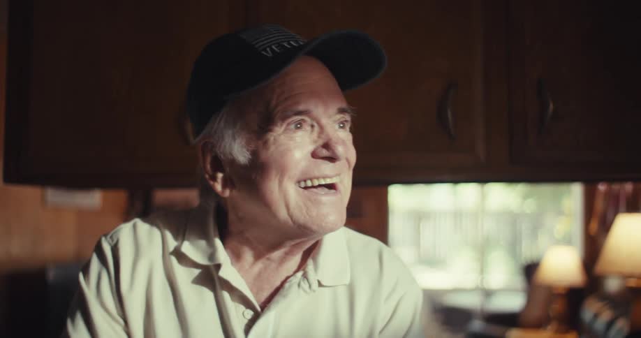 An elderly man inside his kitchen waves at a neighbor outside through his window then begins scrolling through his phone at midday