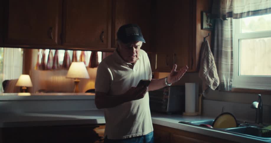 An elderly man standing in the kitchen at midday holding his phone talking and complaining while wearing a hat