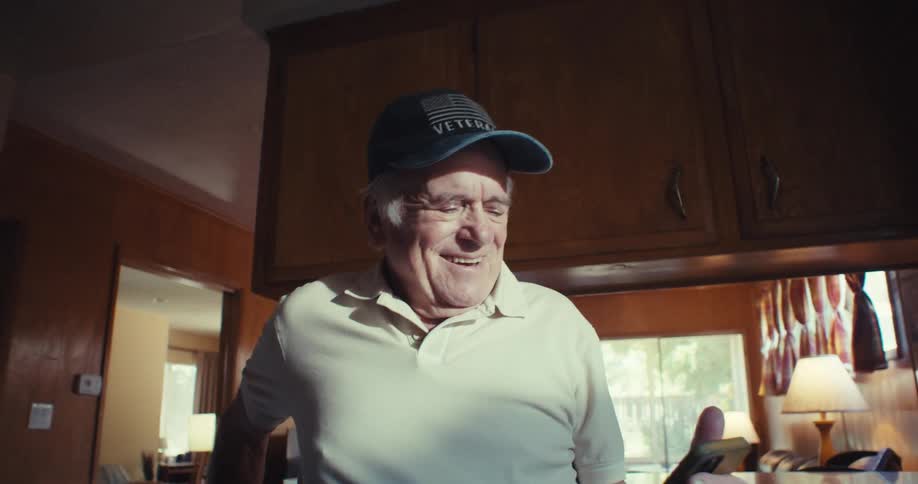 An elderly man smiles while holding a smartphone in a warmly lit kitchen