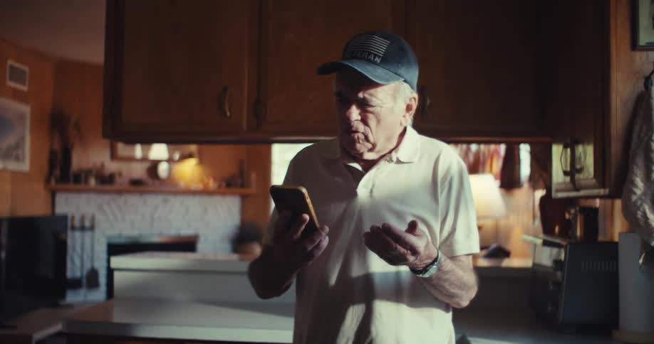 Elderly man in a cap walking around the kitchen and talking on his smartphone