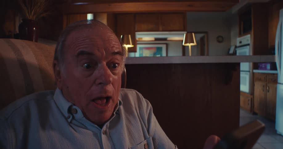 Elderly man making multiple faces while holding a smartphone sitting in an armchair