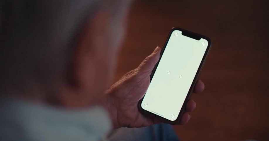 The video shows a closeup of an elderly persons hands holding a smartphone with a green screen as they interact with the touch screen the background is blurred with warm lighting suggesting an indoor setting