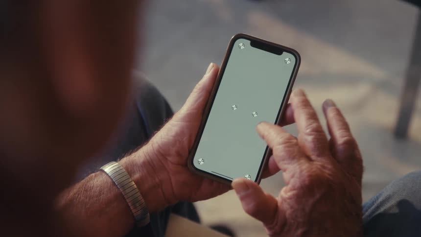 An elderly man sitting indoors scrolling through his smartphone with a focused expression