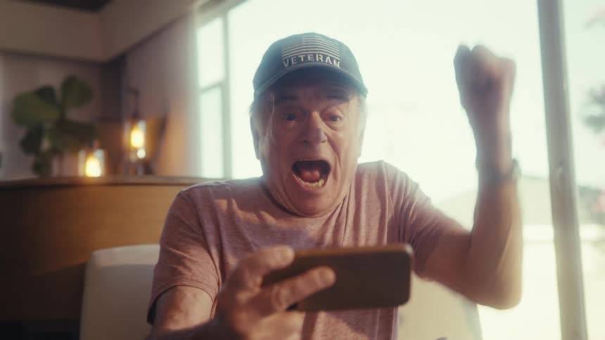 Elderly man raising his hand up and cheering happily while holding a smartphone