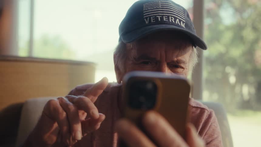 The video shows a closeup of an elderly man wearing a veteran baseball cap and a lightcolored shirt he is holding a smartphone and appears to be waving or interacting with someone on a video call