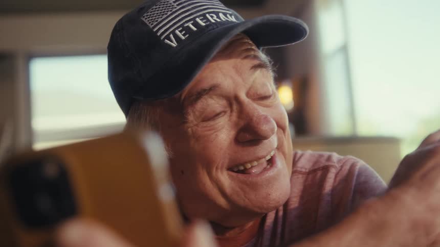 The video features a smiling elderly man wearing a veteran baseball cap holding a smartphone he is looking at the phone and appears to be reacting happily to something on the screen or in a conversation