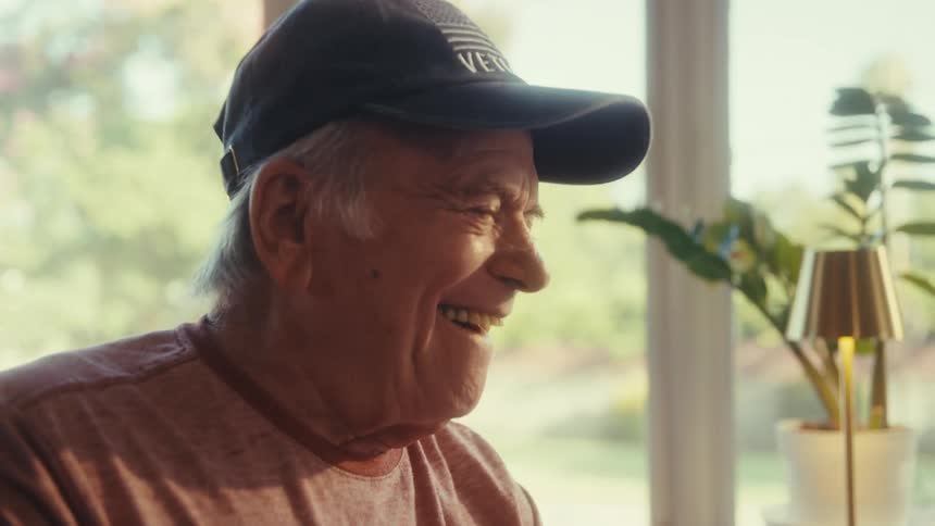 An older man wearing a dark baseball cap and a pink tee shirt smiles and laughs while looking at his cell phone in a brightly lit room