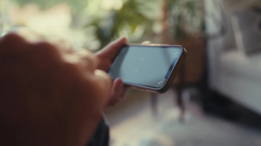 The video shows a persons hands holding a smartphone with a blank reflective screen the background is a brightly lit indoor space possibly a living room with natural light coming from windows