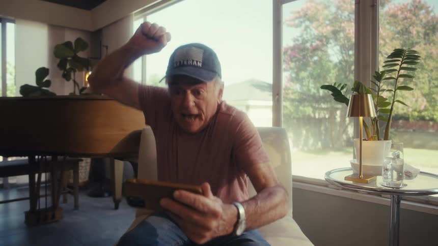 The video features an elderly man wearing a veteran baseball cap and a pink tshirt sitting in a chair and looking at his smartphone he gestures excitedly with his hands indicating he is reacting enthusiastically to something on his phone