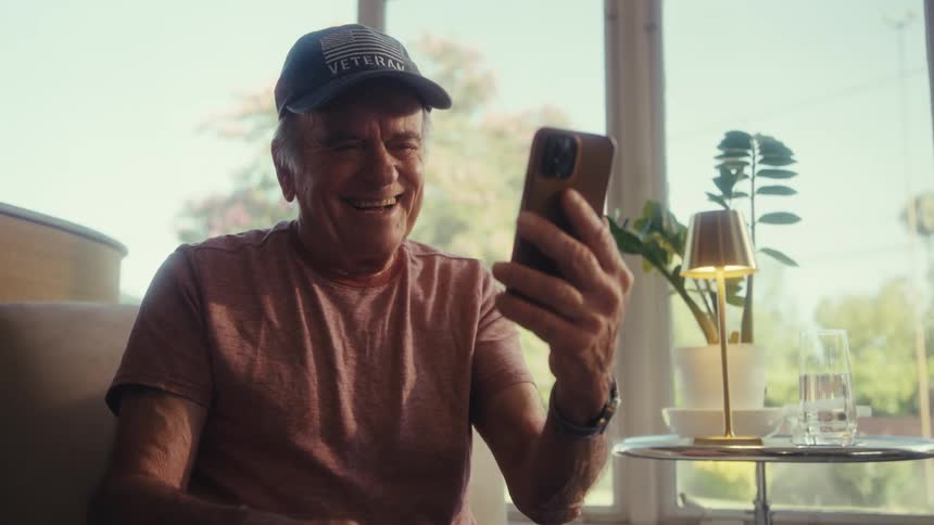 Elderly man wearing a cap sitting interacting with a smartphone and with a happy expression