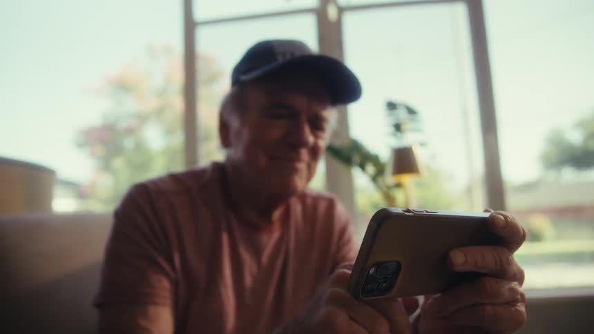 An elderly man wearing a hat is sitting on a couch in his living room at midday operating his smartphone