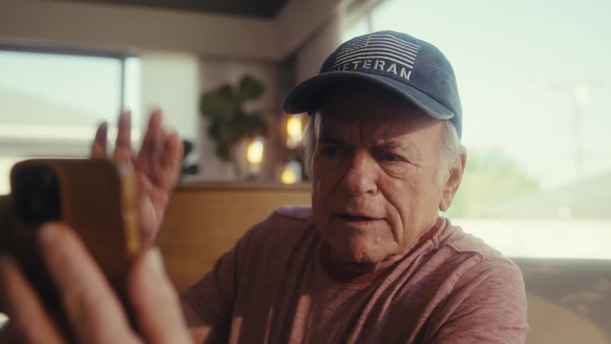 Elderly man sitting and wearing a cap talking happily in front of a smartphone