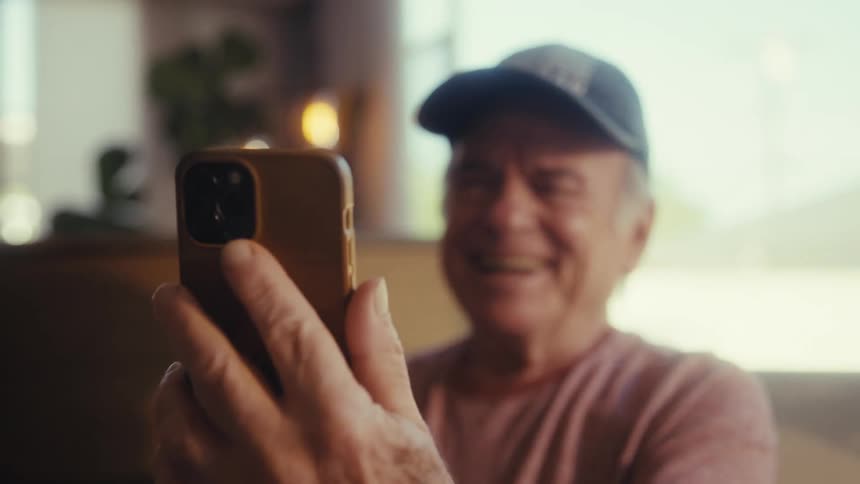 An older man with light colored hair and a light blue collared shirt smiles broadly while talking on his cell phone