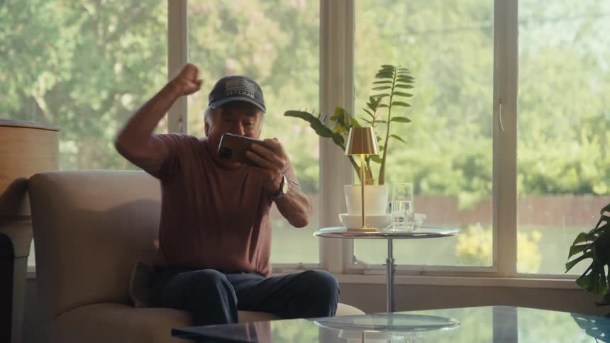 Elderly man bounces on a chair in excitement while watching an event on his smartphone