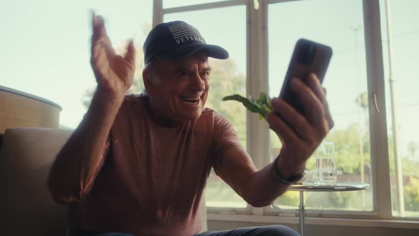 Elderly man waves at phone during video call in cafe