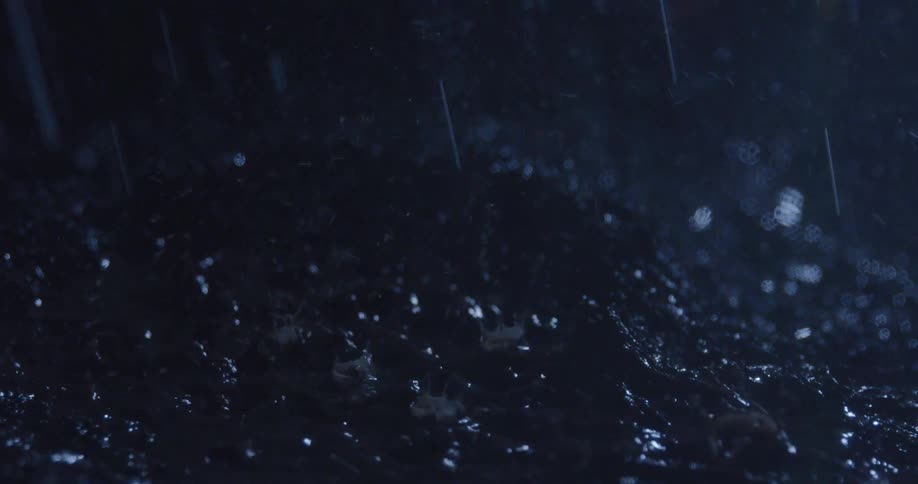 Raindrops splash on a dark wet soil creating shimmering reflections at night
