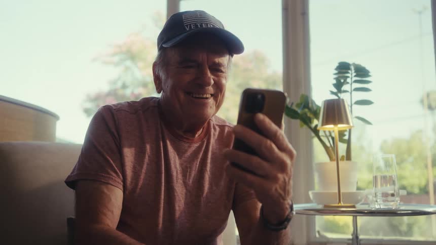 Cheerful elderly man wearing a cap sitting and interacting with a smartphone