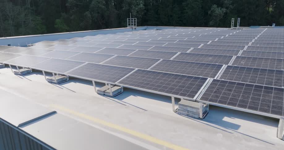 Low approach towards a large modern building surrounded by dense forest with the rooftop covered in rows of solar panels