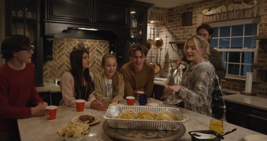 A woman serves food as people enjoy a lively kitchen gathering