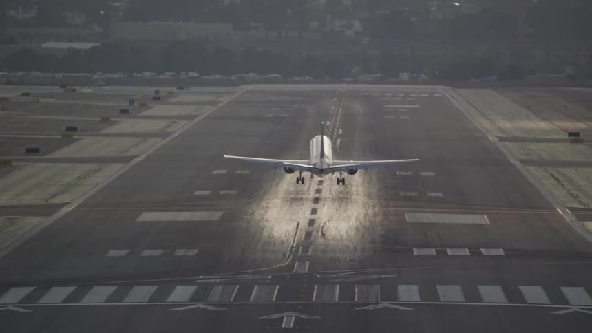 The video shows a wide shot of an airport runway with bright sunlight reflecting off its surface an airplane is seen in the distance approaching for landing