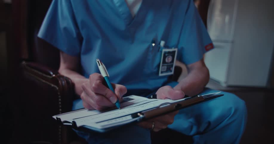 Older male medical professional sits in a living room during a home healthcare visit and fills out a form on a clipboard