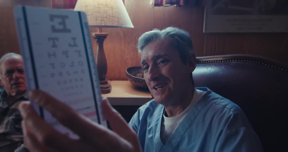 The video shows a man with short gray hair and a blue medical scrub top holding up an eye chart in front of him he appears to be reading or looking at the chart possibly during an eye exam