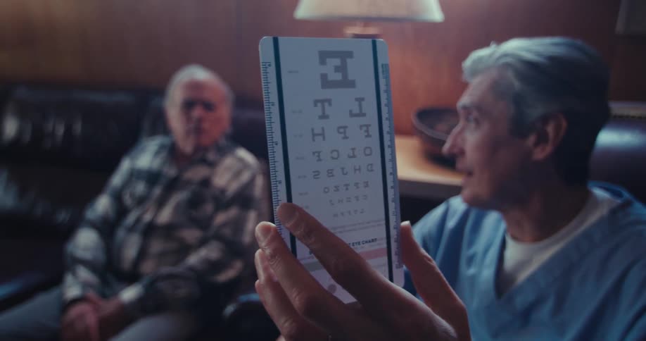 A man in medical attire holds an eye chart in front of an elderly man seated on a couch in a wood paneled room with a lamp