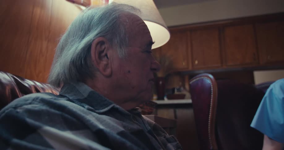 A man in scrubs is talking to an elderly man while both sit in a living room