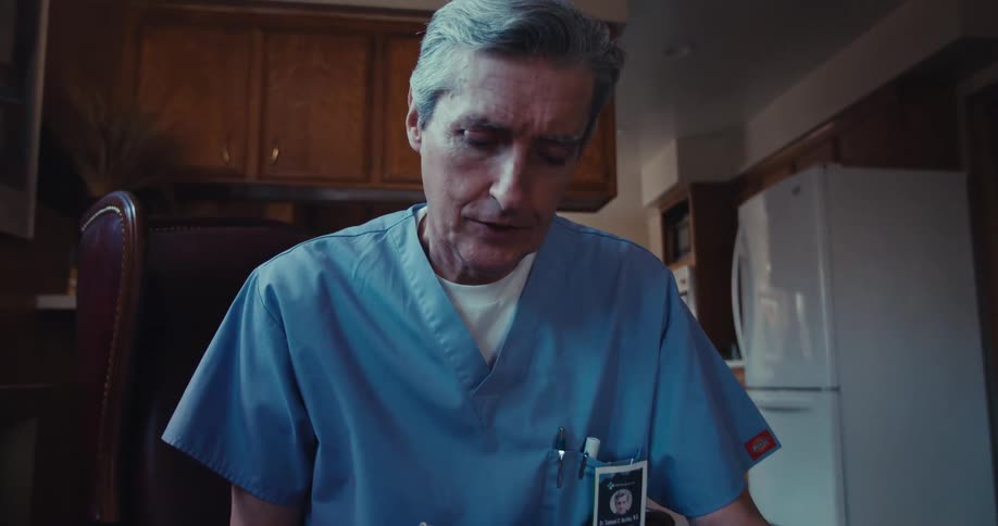 A man in medical scrubs takes notes in a cozy kitchen setting talking to someone not seen
