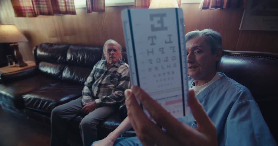 A doctor holds up an eye chart for an older male patient sitting on a couch checking his vision