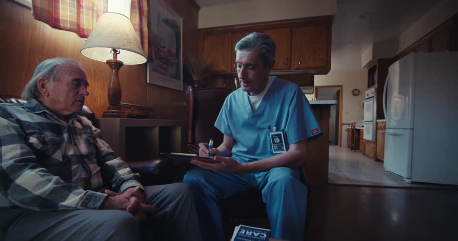 A caregiver consults with the elderly man in a cozy home environment taking notes during the visit