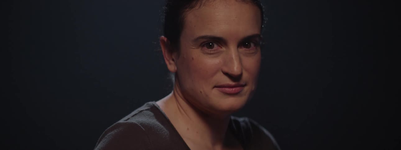 A woman with a neutral expression looks directly at the camera in a dark softly lit setting