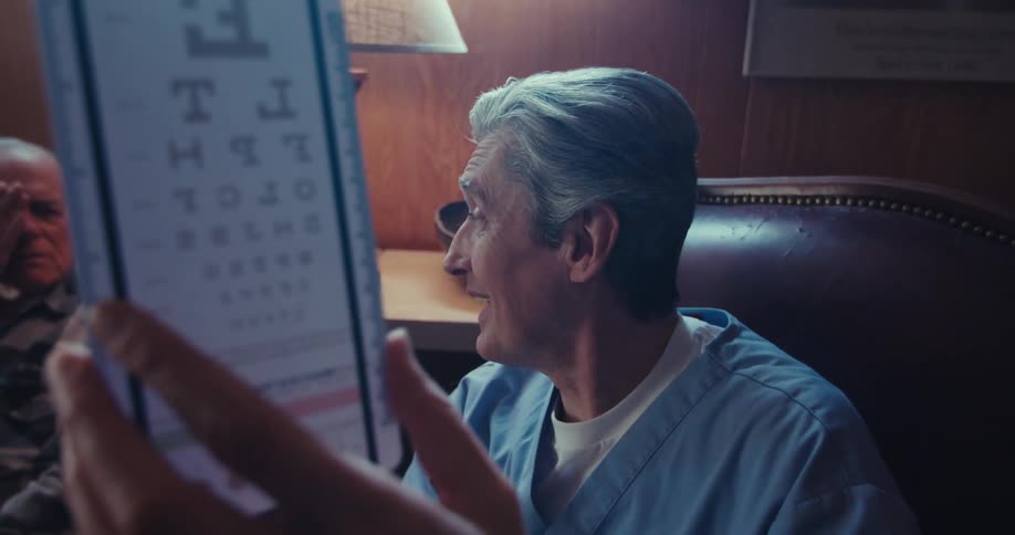The video shows a man in a blue medical scrub top holding up an eye chart he is looking past the chart and appears to be speaking or interacting with someone possibly a patient who is partially visible in the blurred background