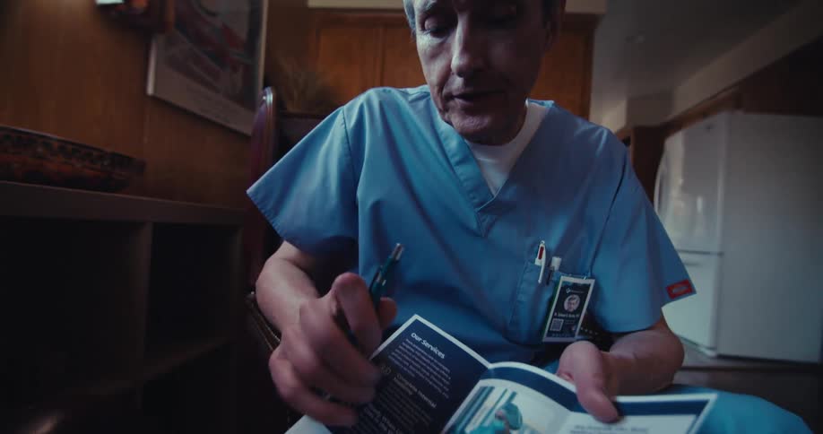 The video shows a man with gray hair wearing a blue medical scrub top looking down and appearing to be speaking his hands are in the foreground and he holds something small possibly a pen or an object he is examining