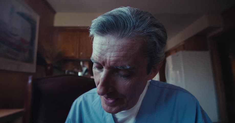 The video shows a person in blue medical scrubs holding a pen and writing on a medical form attached to a clipboard the camera then shifts to a closeup of the persons face revealing gray hair and a focused expression as they continue writing