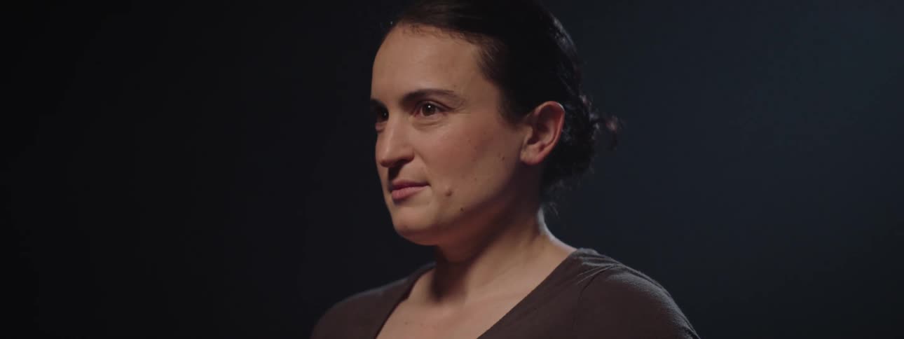 A woman with a prominent arm tattoo and dark hair tied back is shown from the chest up turning her head from left to right as if observing something with light subtly revealing and then obscuring her features against a dark background