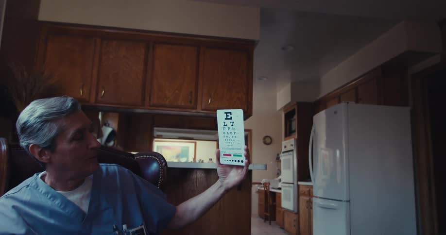 An eye chart is held up by a man in scrubs for an elderly man to read in a living room setting for a vision test
