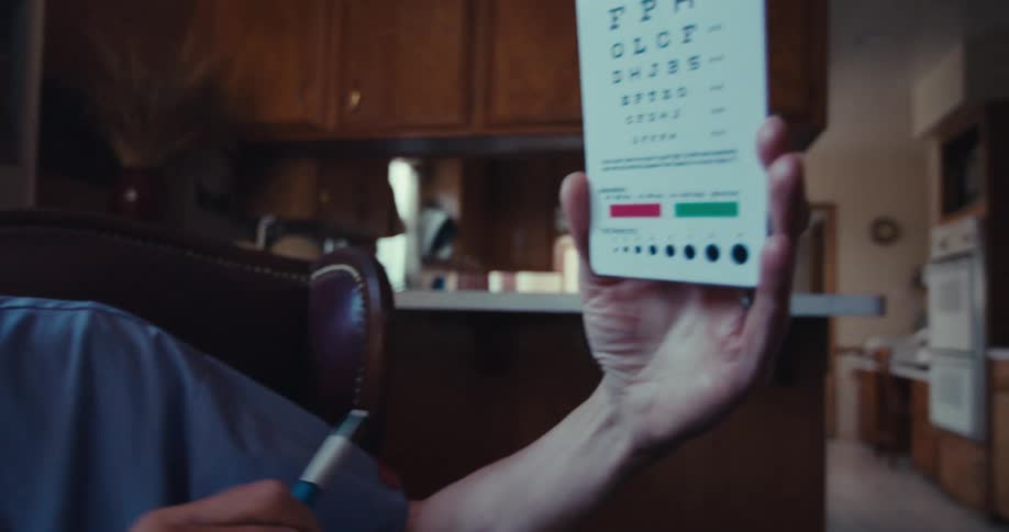 A person in blue scrubs holds up an eye chart while an older man with gray hair sits on a brown leather couch in the background