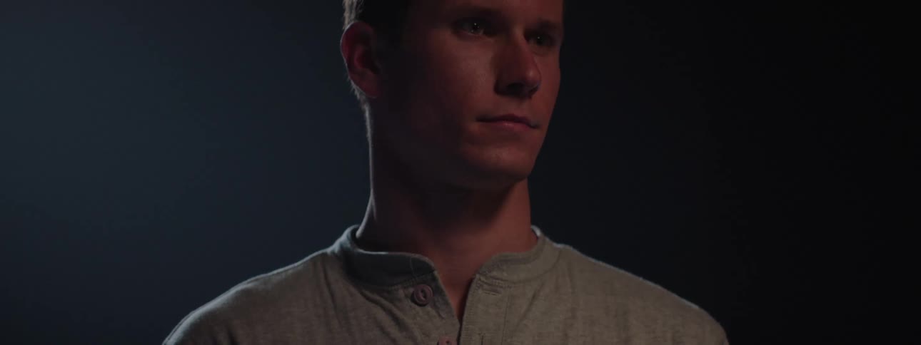 A young man stands on a stage gradually revealed by a slowly intensifying spotlight