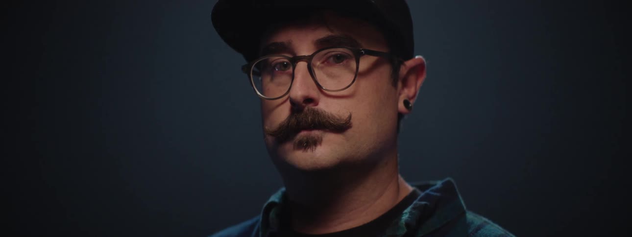 A person with a prominent mustache glasses and a backward cap turns their head to face the camera with light slowly illuminating their face before fading back to darkness