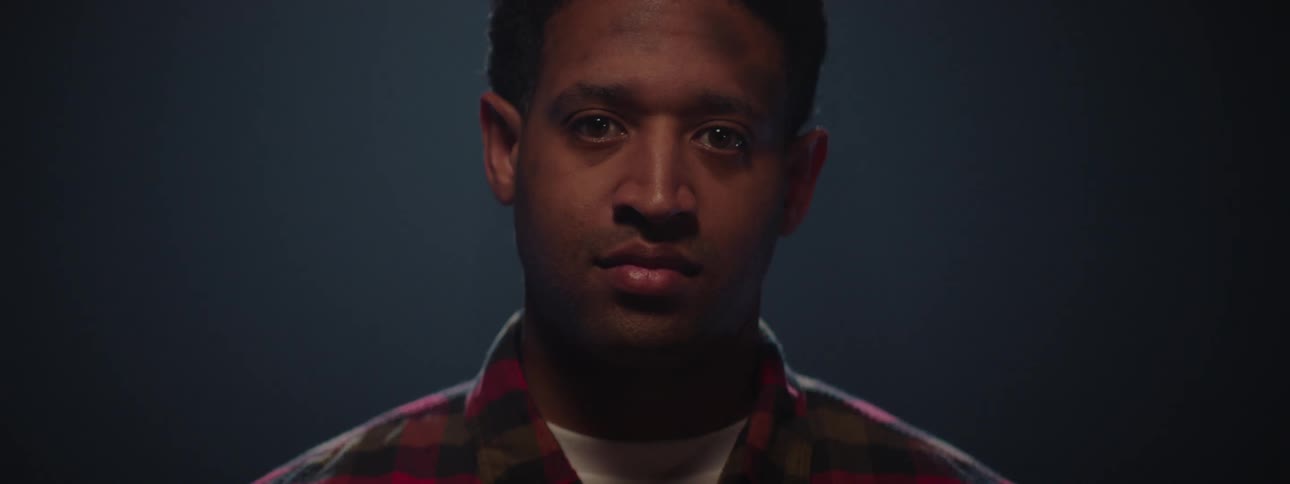 A man with tears in his eyes looks directly into the camera as light slowly reveals his face in a dark setting