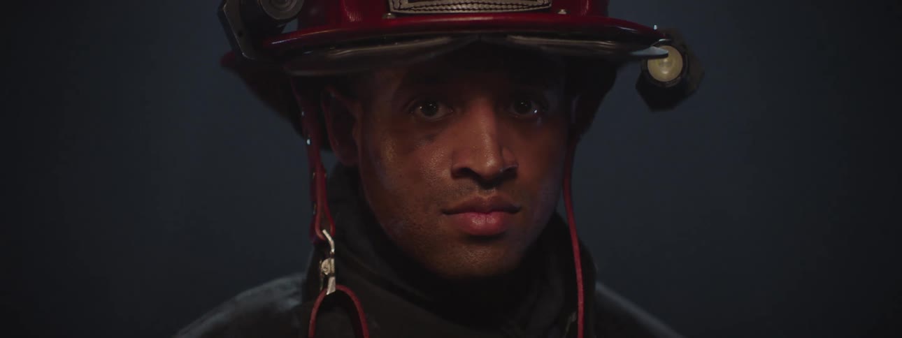 A firefighter wearing a red helmet looks down with a somber expression