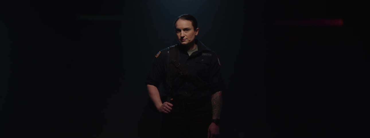 A firefighter looks down solemnly while wearing a red helmet and uniform in a dark area